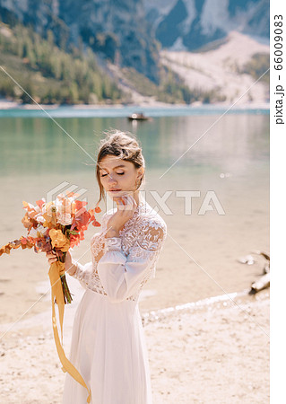 Beautiful bride in a white dress with sleeves and lace, with a yellow autumn bouquet of dried flowers and peony roses, on the Lago di Braies in Italy. Destination wedding in Europe, on Braies lake. 66009083