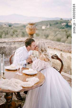 Wedding at an old winery villa in Tuscany, Italy. The wedding couple sits at the dinner table on the roof of the villa and kisses. 66009084