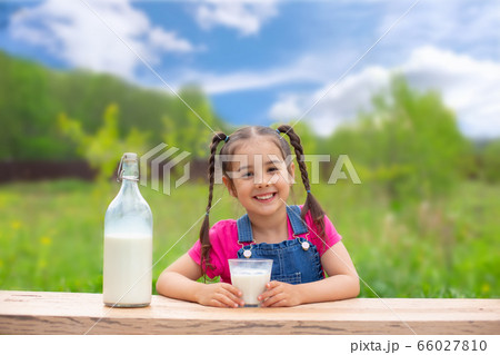 Beautiful little brunette girl sits at a table, holds a glass of milk and smiles, 66027810