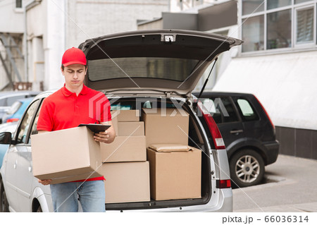Courier holds big box and checks address on tablet, standing by open trunk of car Courier holds big box and checks address on tablet, standing by open trunk of car 66036314