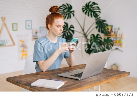 Serious pretty redhead young woman uses the phone while sitting at the table with her laptop. 66037273