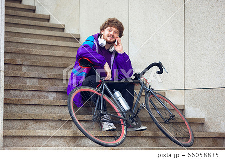 Handsome young man using mobile phone and headphones while sitting near his bicycle beside him Handsome young man using mobile phone and headphones while sitting near his bicycle beside him 66058835