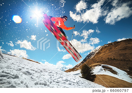 A young stylish man in sunglasses and a cap performs a trick in jumping with a kicker of snow against the blue sky and the sun on a sunny day. The concept of park skiing in winter or spring 66060797