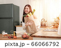 Cute asian woman holding shopping paper bag with fruits and vegetables stand in the kitchen,her look at camera with brightly smile. 66066792