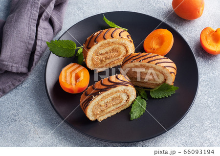 Pieces of sweet roll with cream filling on a plate. Fresh apricot berries and mint leaves. 66093194