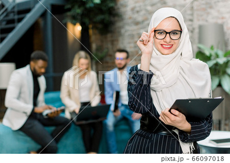 Young pretty Muslim business woman in white hijab and eyeglasses, standing in modern office, holding folder with papers and smiling to camera. Three multiethnic office workers on the background 66097018