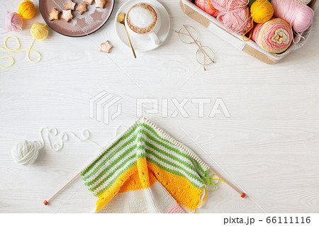Knitted bright striped fabric. Woolen and cotton yarn lies in a basket. Cappuccino coffee, cookies and glasses lie on a white wooden background. Knitted bright striped fabric. Woolen and cotton yarn lies in a basket. Cappuccino coffee, cookies and glasses lie on a white wooden background. 66111116