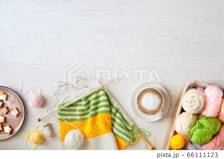 Knitted bright striped fabric. Woolen and cotton yarn lies in a basket. Cappuccino coffee, cookies and glasses lie on a white wooden background. 66111123
