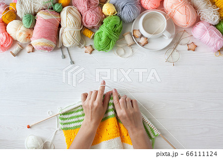 Hands of craftswoman knit a bright knitted striped fabric. Woolen and cotton yarn lies in a basket. Cappuccino coffee, cookies and glasses lie on a white wooden background. 66111124
