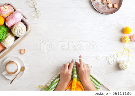 Hands of craftswoman knit a bright knitted striped fabric. Woolen and cotton yarn lies in a basket. Cappuccino coffee, cookies and glasses lie on a white wooden background. 66111130