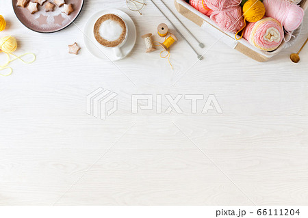 Woolen and cotton bright yarn lies in a basket. Cappuccino coffee, cookies and glasses lie on a white wooden background. 66111204