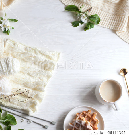 Knitting, knitting needles, yarn, coffee and waffles stand on a white wooden background. Surrounded by branches of a blossoming apple tree. Flatlay 66111305