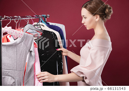 Portrait of an attractive girl on a red background among things hanging on a hanger. Concept Nothing to wear. Woman decides what to wear. Fashion style 66131543