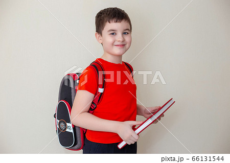 A smiling schoolboy in a red T-shirt, with a satchel behind his back, stands and holds a big book, looks at the camera A smiling schoolboy in a red T-shirt, with a satchel behind his back, stands and holds a big book, looks at the camera 66131544
