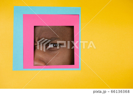 Eye of african-american man peeking throught square in yellow background Eye of african-american man peeking throught square in yellow background 66136368