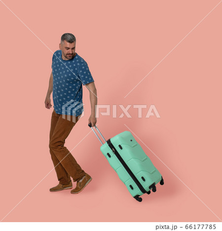 Man with green plastic suitcase posing in studio on pink background. Elegance Caucasian man in blue t-shirt and brown trousers goes on vacation. Travel concept. Weekend concept 66177785