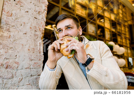 Side angle view of young handsome hungry man eating croissant while sitting in outdoor cafe. Portrait of handsome young Caucasian man eating delicious croissant Side angle view of young handsome hungry man eating croissant while sitting in outdoor cafe. Portrait of handsome young Caucasian man eating delicious croissant 66181956