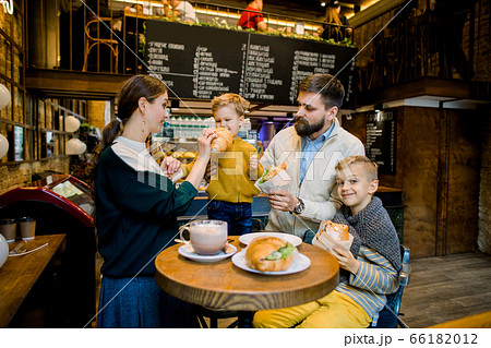 Young cheerful family, bearded father, pretty mother and two children boys, drinking hot chocolate drink and eating traditional French croissants in a cozy indoor city cafe. 66182012