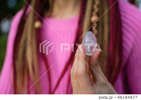 Adult girl in knitted sweater with long dreadlocks is holding transparent violet amethyst yoni egg for vumfit, imbuilding or meditation outdoors on her body background outdoors Adult girl in knitted sweater with long dreadlocks is holding transparent violet amethyst yoni egg for vumfit, imbuilding or meditation outdoors on her body background outdoors 66184877