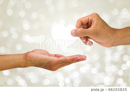 Hand giving white heart to another hand on blurred white glittering bokeh background 66203820