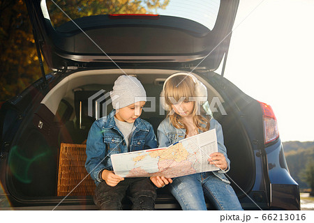 Agreeable boy and girl are looking at the road map while sitting in the auto's trunk and discussing the move direction. Family vacation trip by car. 66213016