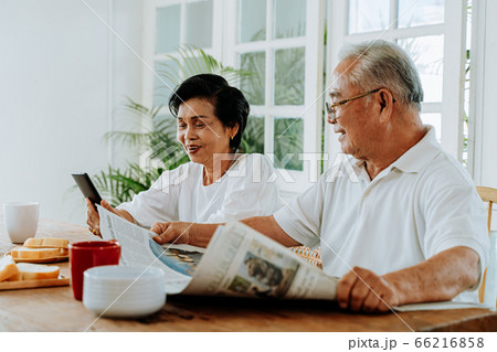 Senior Asian couple having breakfast together in dinner room. 70s retired elderly man reading newspaper while woman using mobile phone. Senior Asian couple having breakfast together in dinner room. 70s retired elderly man reading newspaper while woman using mobile phone. 66216858