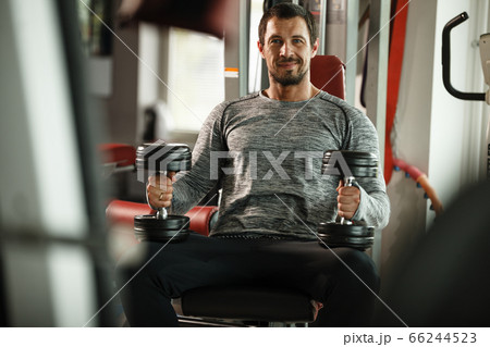 Fitness in gym, sport and healthy lifestyle concept. Handsome athletic man in grey shirt making exercises. Bodybuilder male model training biceps muscles with dumbbells 66244523