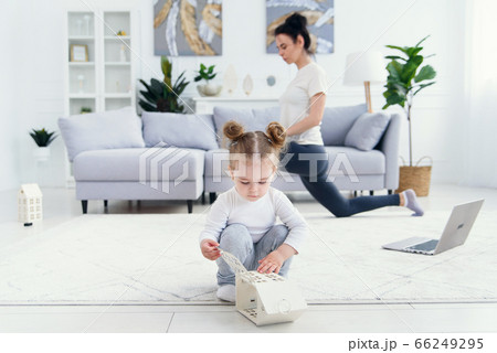 Funny baby girl playing at home while her sporty healthy mommy doing fitness and yoga exercises on the background. 66249295