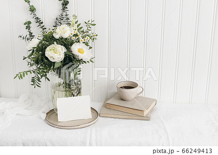 Feminine wedding or birthday table mockup scene with floral bouquet. White peonies, tanacetum flowers, eucalyptus, greeting card,cup of coffee and books. White wooden wall background. Rustic interior. 66249413