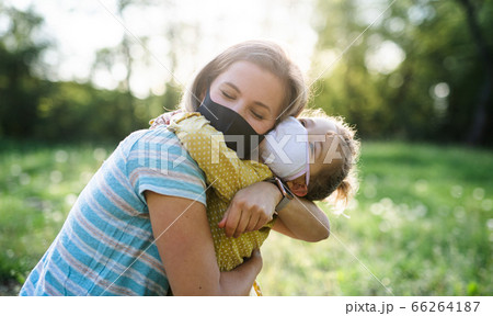 Side view of happy mother hugging small daughter in nature, wearing face masks. Side view of happy mother hugging small daughter in nature, wearing face masks. 66264187