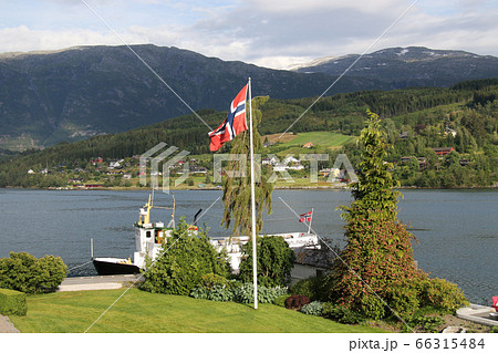 ノルウェー　ハダンゲルフィヨルド、ウルヴィク地区に停泊中の遊覧船とノルウェーの国旗 66315484
