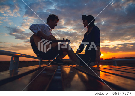 Silhouette of a man with a guitar and a woman in a hat sitting on the roof of a car on the background of the sunset. Silhouette romance concept. 66347577