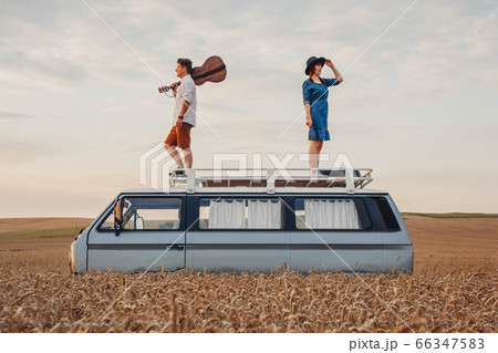Young couple man with a guitar and woman in a hat are standing on the roof of a car in a wheat field. Travel and adventur 66347583