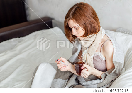 A sick young woman, sitting in bed with a scarf, suffers from a sore throat. Drinking hot tea. A woman with a fever wrapped in a blanket. A sick young woman, sitting in bed with a scarf, suffers from a sore throat. Drinking hot tea. A woman with a fever wrapped in a blanket. 66359213