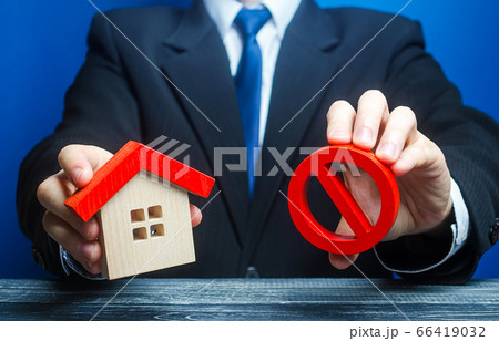 A man holds a house and the red prohibition symbol NO. Bank refuses to provide a mortgage loan. Housing problems. Confiscation pledged property. Prohibition of operation. Denial of insurance payment. 66419032