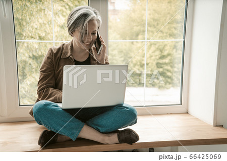 Mature freelance woman talks mobile phone working laptop computer while sitting tailor-fashion on wooden silll. Tinted image Mature freelance woman talks mobile phone working laptop computer while sitting tailor-fashion on wooden silll. Tinted image 66425069