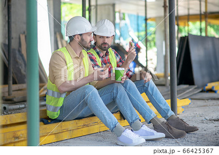 Building workers in yellow vests and helmets sitting on boards, having coffee with sandwiches, discussing something 66425367