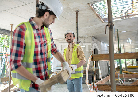 Building workers in yellow vests and gloves carrying together wooden board Building workers in yellow vests and gloves carrying together wooden board 66425380