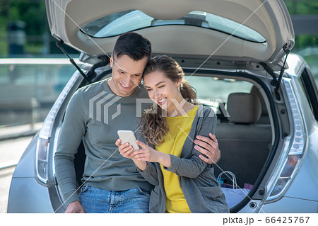 Woman with smartphone and man crouched on car trunk. Woman with smartphone and man crouched on car trunk. 66425767