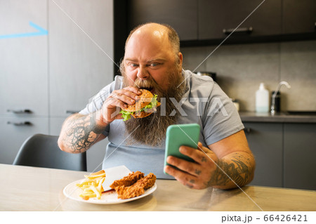 Bearded tattooed involved man with smartphone and burger. 66426421