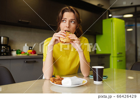 Dark-haired girl biting off a burger looking to the side 66426429