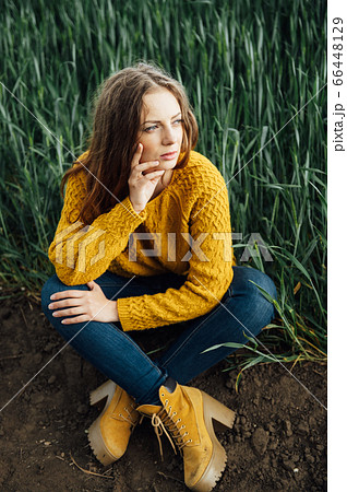 Beautiful girl with jeans, a yellow sweater and boots sits near an early wheat field 66448129