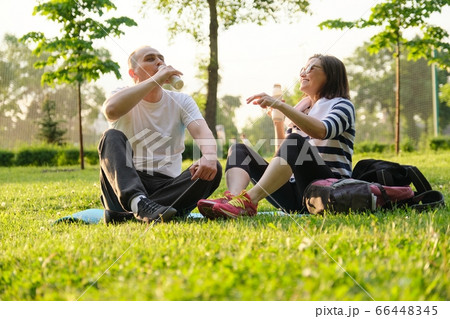 Happy mature couple sitting in the park on fitness mat, resting drinking yogurt after sports exercises. 66448345