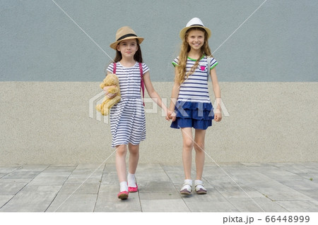 Two little beautiful girlfriends holding hands, girls walking in striped dresses, hats with backpack 66448999