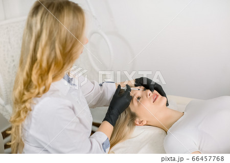 Young pretty blond woman receiving treatments in the beautician office lying on the couch. Young beautiful woman cosmetologist making injections into patient face. Contouring procedure, revitalization Young pretty blond woman receiving treatments in the beautician office lying on the couch. Young beautiful woman cosmetologist making injections into patient face. Contouring procedure, revitalization 66457768