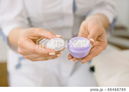 Cropped image of woman doctor cosmetologist with bowl of natural cream, closeup. Cosmetologist's hands holding peeling cream in small violet box. Focus on the box with cream Cropped image of woman doctor cosmetologist with bowl of natural cream, closeup. Cosmetologist's hands holding peeling cream in small violet box. Focus on the box with cream 66488789