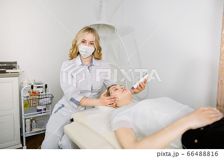 Pretty young woman cosmetologist posing to camera while making laser therapy for her female client lying on the couch. Treatment of woman at beautician office. Hardware cosmetology Pretty young woman cosmetologist posing to camera while making laser therapy for her female client lying on the couch. Treatment of woman at beautician office. Hardware cosmetology 66488816