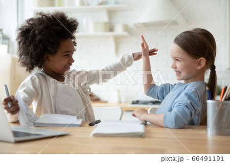 Multiethnic girls distracted from homework playing seated at table indoor Multiethnic girls distracted from homework playing seated at table indoor 66491191