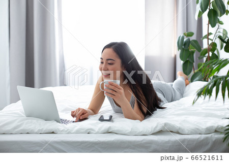 Happy female with tea using notebook on bed 66521611