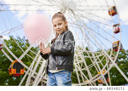 Portrait of a child with sweet cotton candy. A little girl on the background of ferris wheel is eating candy-floss summer sunny day. 66548151